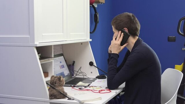 Young woman answers the phone while she is in smart working at home in the room, sitting at the desk in front of the laptop