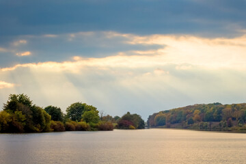 Sunlight penetrates the dark clouds over the river, the river and the forest in the distance with a picturesque sky
