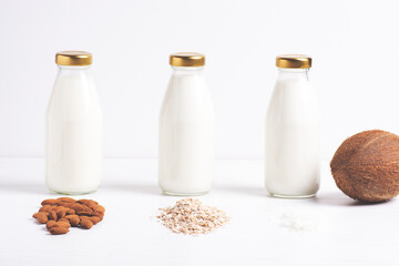 Three glass bottles with vegetarian milk stand on a white table, next to them are almonds, oatmeal and coconut. The concept of diet food, dairy-free products, horizontal orientation.