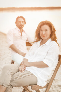 Beautiful mature couple resting on the seashore. Selective focus on red-haired woman in white clothes looking at the camera in the foreground. Toned image.