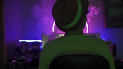 Rear view of resting man smoking a hookah at home workplace. Man in hat relaxes in a dark room with...