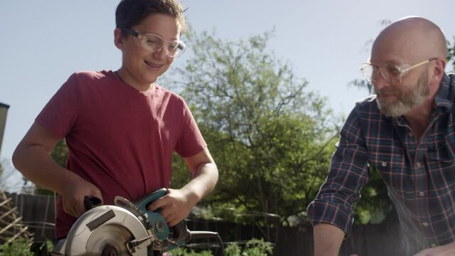 Proud father watches as his teenage son cuts a piece of wood with a circular saw, they high five