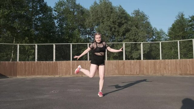 young woman dancer in a mesh T-shirt and black short shorts dancing street modern dance, twerking, shaking her buttocks against the background of houses and green trees. Slow motion. Gimbal shot