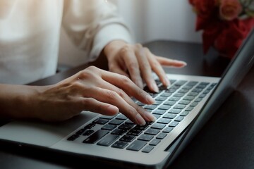 Hands of Asian woman typing report, presentation or operations of internet banking on keyboard of laptop on black desk with red flowers 