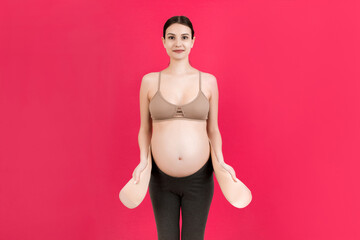 Portrait of pregnant woman dressing maternity belt against pain in the back at pink background with copy space. Orthopedic abdominal support belt concept