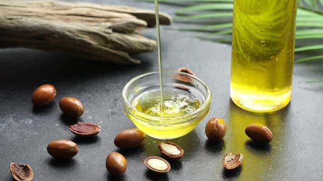 Argan oil pouring into a glass bowl with argan seeds on a dark background in sunlight. Argan oil based cosmetics concept.