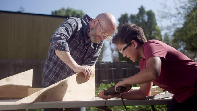 A father helps his son as he drives a screw into a wood construction project with a power drill 