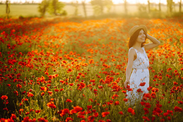 Beautiful girl in the poppy field. Portrait of a cute brunette girl in poppy field at sunset in a white dress and hat. Summertime.