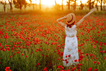 Beautiful girl in the poppy field. Portrait of a cute brunette girl in poppy field at sunset in a white dress and hat. Summertime.
