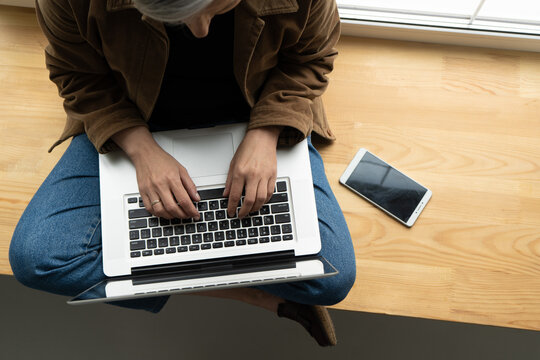 Grey haired businesswoman types at laptop keyboard sitting on wooden windowsill with mobile phone on it. High angle view. Close up shot. Tinted image.