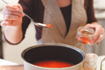 Female cook adds paprika spices while cooking tomato soup.Close-up view on spoon with red pepper.