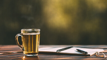 Transparent cup with hot tea with steam and an opened paper notebook with a pen on a textured wooden table