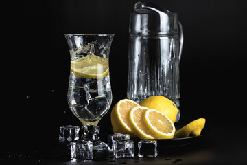 lemon slices fall into a glass on the stem. Carafe of water. Still life of glass objects on a black background. Cold lemonade. Yellow on black.