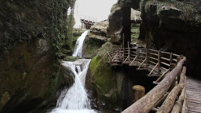 Small waterfalls in a rocky gorge full of vegetation with a wooden walkway, Caglieron Caves, Italy. Video that inspires tranquility and light-heartedness in nature, trips in beautiful natural settings