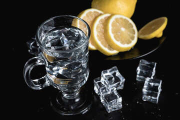 Sliced lemon on a black plate. A decanter and a tall glass of water and ice. Still life of glass objects on a black background. Cold lemonade. Yellow on black.