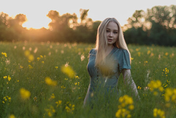 Naklejka na ściany i meble Young happy blonde girl staying at a green wheat field in the evening against the background of the sun shines. Concept of freedom. Summer time joy. Beautiful young woman walking on field.