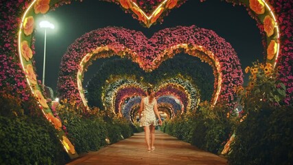 Beautiful garden view evening romantic floral blooming arches in shape hearts orange illumination. Silhouette woman tourist turned away, walks through night flower garden. Back rear view. Dubai UAE 4k