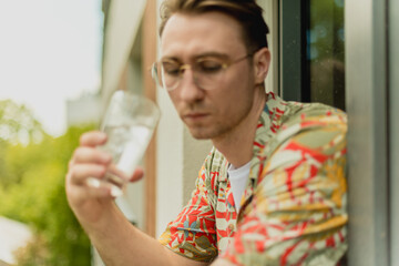 Young healthy man drinking fresh mineral pure ice water holding glass of drink. Thirsty guy standing at balcony looking outside with cocktail. Male hang out concept in summer.