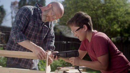 A father and son wearing safety glasses build a wooden skateboard ramp in the backyard