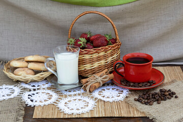 On the table is a basket with strawberries, a cup of coffee, coffee beans, cinnamon sticks, a cup of milk and a plate of cookies.