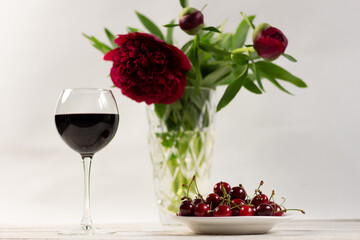 Red wine glass, ripe cherry berries in plate and peon flowers bouquet in vase on white wooden background