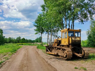 old yellow bulldozer stands on the side of a country road near trees against a blue sky with clouds on a sunny day
