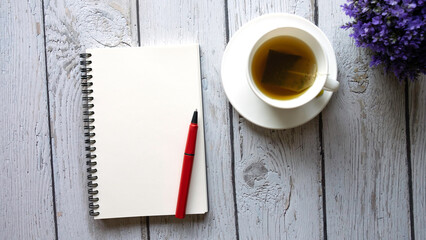 Top view blank open notebook with red pen on wooden table.A cup of hot tea with saucer and purple plant on table.