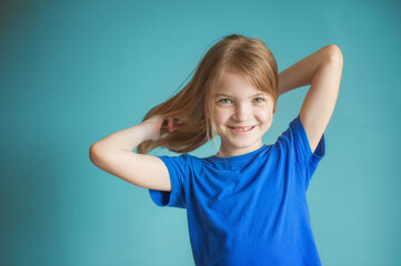 Close-up studio shot of a lovely blond little girl in a blue t-shirt posing against a blue background. Holds hair