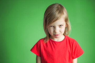 Close-up studio shot of a lovely blond little girl in a red t-shirt and posing against a green background. Offended and looking sullenly