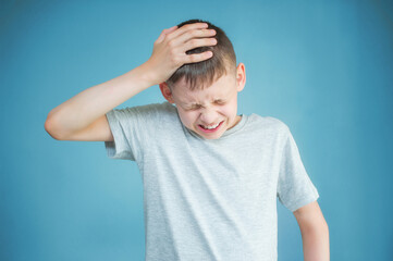 Portrait of a teenager in a gray t-shirt. Photo of a young boy on a blue background. He holds on to his head. The concept of headache, stress, anger, misunderstanding. Copy space