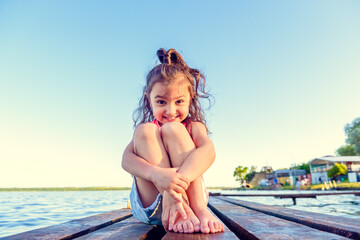 Fototapeta na wymiar little happy girl with curly hair sits on a wooden pier in the summer on the lake. Looking in frame