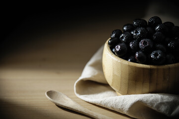Fresh blueberries with small drops of water on the surface, in a bamboo bowl on a wooden table, with a tablecloth below and a wooden spoon.