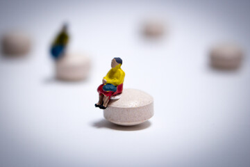 The miniature old woman in yellow outfit sitting on the tablet (pill) in the white background. Many tablets are randomly placed and the woman keeps distance away from the guy.