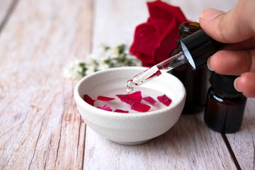 Woman of hand holding dropper and dripping oil into white bowl.Brown bottles,red rose and white cutter flowers on white wooden table.Aromatherapy and essential organic concept.