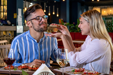 Happy young romantic couple in love feeding each other with bruschetta at dinner in a beautiful fancy restaurant.