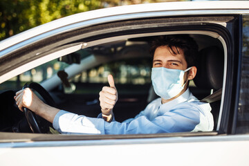 Young taxi driver shows thumb up sign sitting in the car and wearing protective sterile medical mask, works hard during coronavirus outbreak. Social distance, virus spread prevention, treat concept.