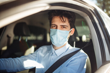 Closeup portrait of young man sits in the car wearing sterile medical mask. Boy taxi driver works hard during coronavirus outbreak. Social distance, virus spread prevention and treat concept.