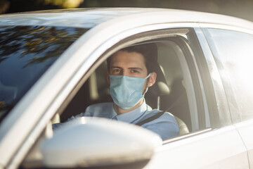 Fototapeta premium Closeup portrait of young man sits in the car wearing sterile medical mask. Boy taxi driver works hard during coronavirus outbreak. Social distance, virus spread prevention and treat concept.