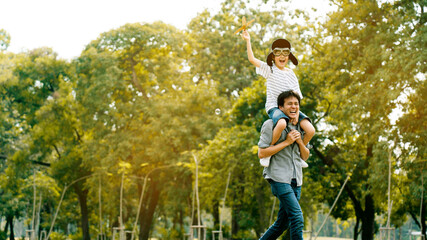 Lifestyle Asian family Father and son sat on his shoulders and run in  park. Paper plane as a toy in hand of kid And there are many big trees in garden Is natural in the morning of summer