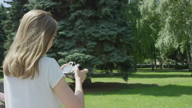 Blonde bespectacled girl assessing taken photos and taking new pictures of sunlit lawn with birch and fir trees in park. View from behind.