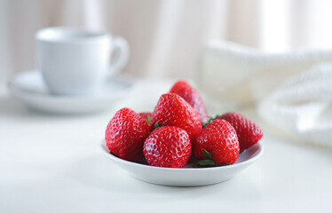 Strawberries in a white plate on a white table, behind is a white mug and a white towel lies.