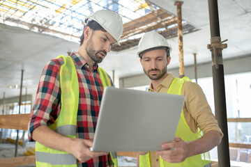 Building workers in yellow vests holding laptop, checking something on it