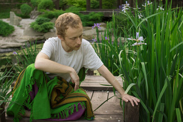 Young handsome man sits on a wooden bridge in the grass and reaches for a flower
