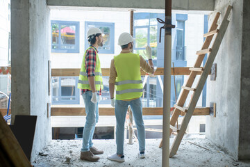 Fototapeta premium Building workers in yellow vests and gloves standing back to camera near the window, discussing something
