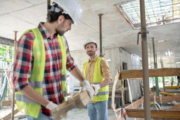 Fototapeta premium Building workers in yellow vests and gloves carrying together wooden board