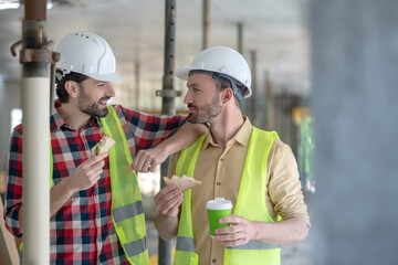Building workers in yellow vests and helmets having lunch, talking smiling