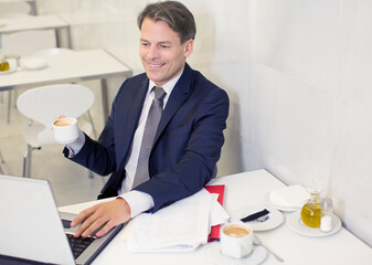 Smiling businessman drinking coffee and using laptop in restaurant