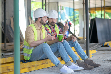 Building workers in yellow vests and helmets sitting on boards, having coffee with sandwiches, discussing something