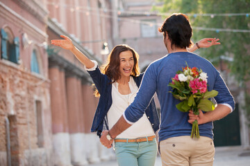 Enthusiastic woman approaching man with flowers behind back