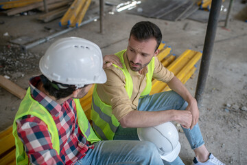 Tired building workers in yellow vests sitting on boards, one of men putting his hand on coworker shoulder